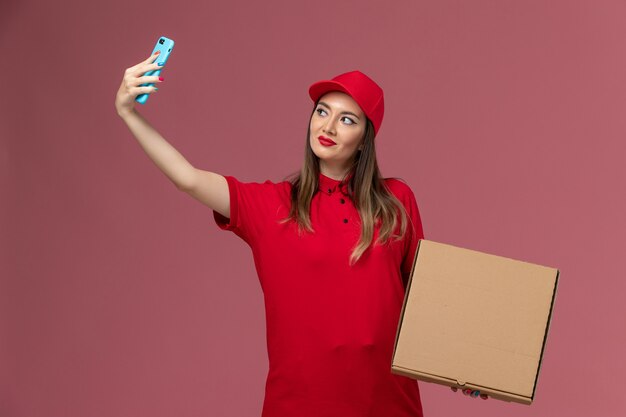 Vista frontal joven mensajero en uniforme rojo sosteniendo la caja de comida de entrega y tomando fotos con ella sobre fondo rosa empresa uniforme de servicio de entrega