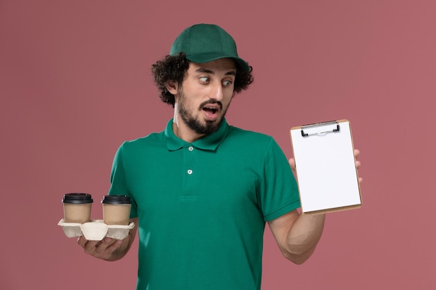 Vista frontal joven mensajero en uniforme verde y capa sosteniendo tazas de café de entrega y bloc de notas sobre fondo rosa servicio de trabajador masculino entrega uniforme