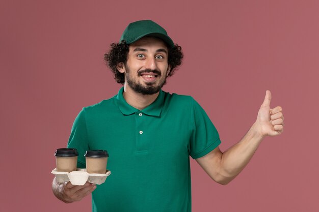 Vista frontal joven mensajero en uniforme verde y capa sosteniendo tazas de café de entrega en la entrega uniforme de trabajo de servicio de fondo rosa