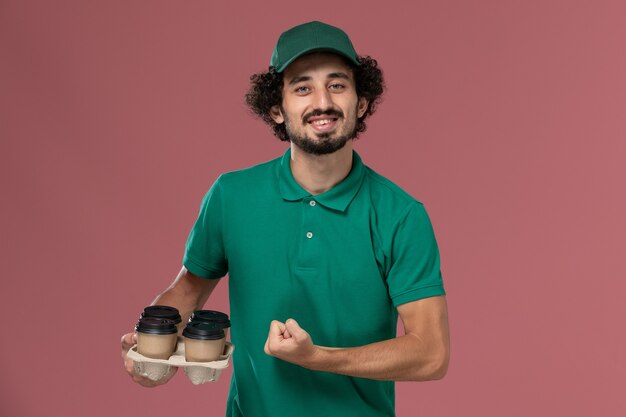 Vista frontal joven mensajero en uniforme verde y capa sosteniendo tazas de café de entrega marrón flexionando en el trabajo de trabajador de entrega uniforme de servicio de fondo rosa