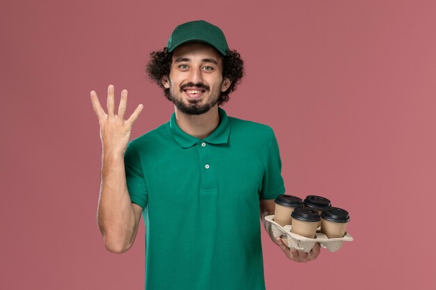 Vista frontal joven mensajero en uniforme verde y capa sosteniendo tazas de café de entrega marrón con sonrisa sobre fondo rosa servicio de entrega uniforme trabajo masculino