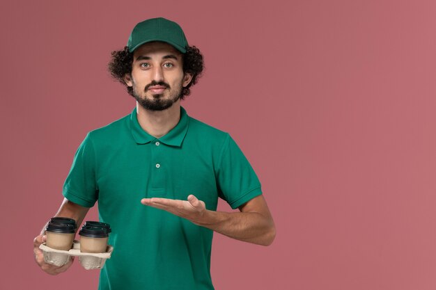 Vista frontal joven mensajero en uniforme verde y capa sosteniendo tazas de café de entrega marrón en el trabajo de trabajador de entrega uniforme de servicio de fondo rosa claro