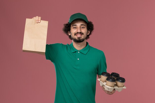 Vista frontal joven mensajero en uniforme verde y capa sosteniendo tazas de café de entrega con paquete de comida sobre fondo rosa servicio entrega uniforme