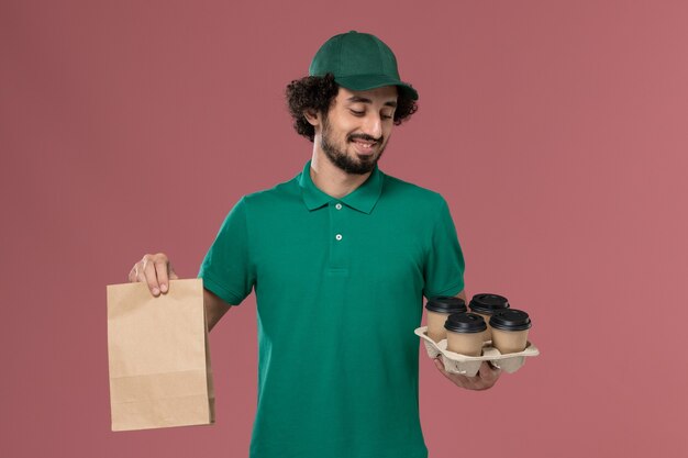 Vista frontal joven mensajero en uniforme verde y capa sosteniendo tazas de café de entrega con paquete de comida en el trabajo de entrega uniforme de servicio de fondo rosa