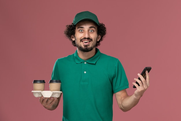 Vista frontal joven mensajero en uniforme verde y capa sosteniendo tazas de café de entrega y su teléfono con sonrisa en servicio de escritorio rosa entrega uniforme de trabajo