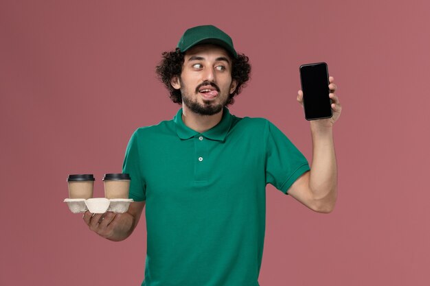 Vista frontal joven mensajero en uniforme verde y capa sosteniendo tazas de café de entrega y su teléfono en el trabajador de entrega uniforme de trabajo de servicio de fondo rosa