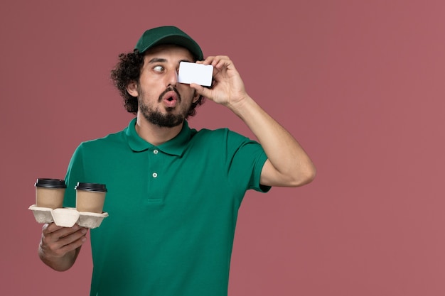Vista frontal joven mensajero en uniforme verde y capa sosteniendo tazas de café de entrega y tarjeta sobre fondo rosa servicio trabajo entrega uniforme masculino