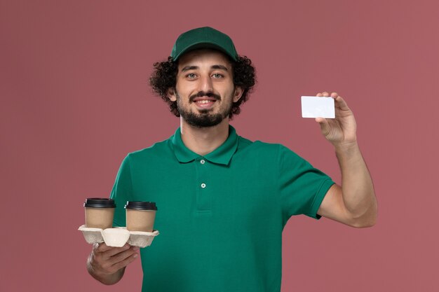 Vista frontal joven mensajero en uniforme verde y capa sosteniendo tazas de café de entrega y tarjeta sobre fondo rosa servicio trabajo entrega uniforme