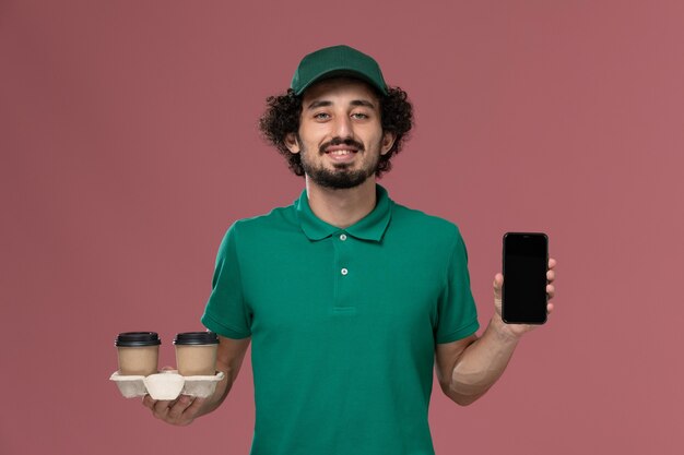 Vista frontal joven mensajero en uniforme verde y capa sosteniendo tazas de café de entrega y teléfono en la entrega uniforme de trabajo de servicio de fondo rosa claro