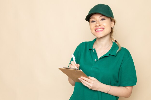 Vista frontal joven mensajero en uniforme verde y capa verde anotando notas sonriendo en el uniforme de mujer de espacio ligero