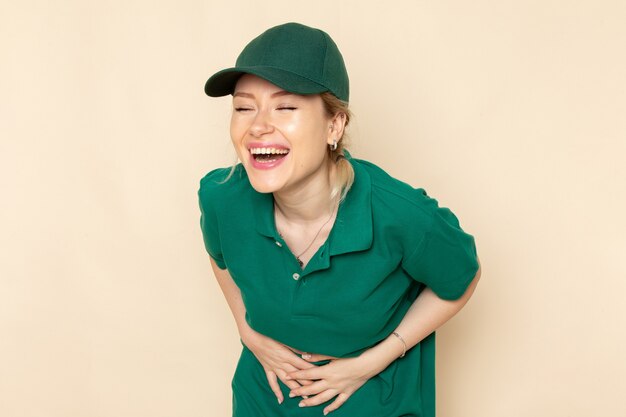 Vista frontal joven mensajero en uniforme verde y capa verde riendo en el espacio de luz uniforme de trabajo de mujer