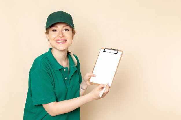 Vista frontal joven mensajero en uniforme verde y capa verde sosteniendo el bloc de notas sonriendo en el trabajo de espacio ligero