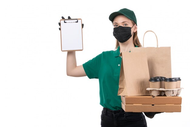 Una vista frontal joven mensajero en uniforme verde guantes negros y máscara negra sosteniendo paquetes de entrega de alimentos entregando