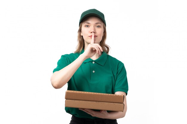 Una vista frontal joven mensajero en uniforme verde sonriendo sosteniendo cajas de comida mostrando signo de silencio