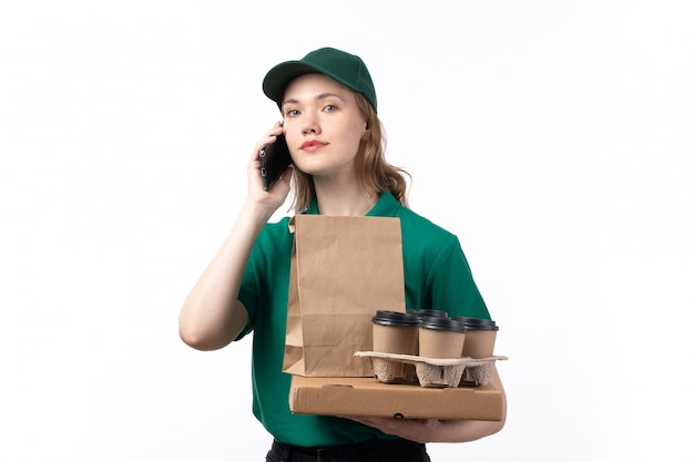 Una vista frontal joven mensajero en uniforme verde sonriendo sosteniendo tazas de café paquetes de alimentos hablando por teléfono