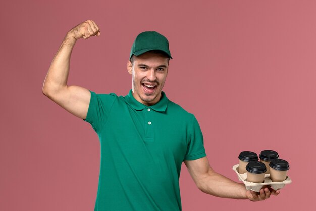 Vista frontal joven mensajero en uniforme verde sosteniendo tazas de café marrón y flexionando en servicio de fondo rosa claro