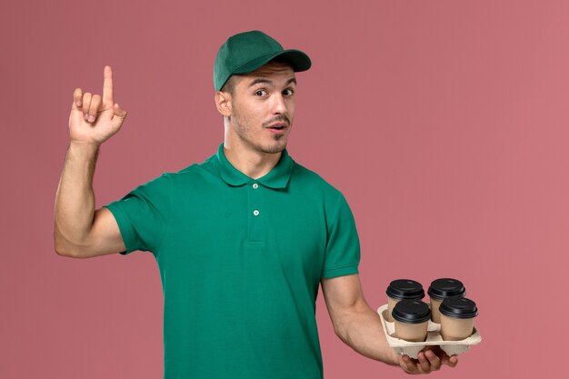 Vista frontal joven mensajero en uniforme verde sosteniendo tazas de café marrón levantando el dedo sobre el piso rosa claro