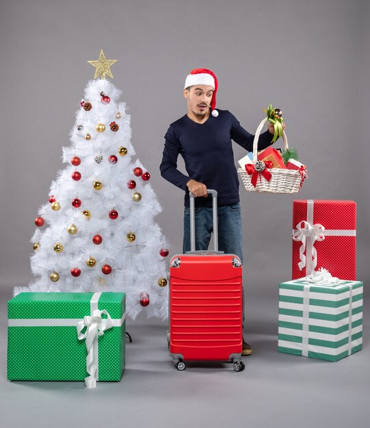 Vista frontal joven mirando la canasta de regalo de pie cerca del árbol de Navidad blanco y coloridos regalos sobre fondo gris aislado