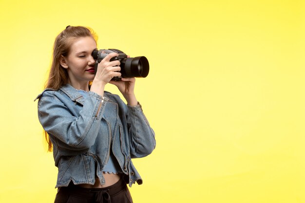 Una vista frontal joven moderna en camisa azul pantalón negro y abrigo de jean posando feliz expresión sonriente sosteniendo la cámara de fotos tomando fotos