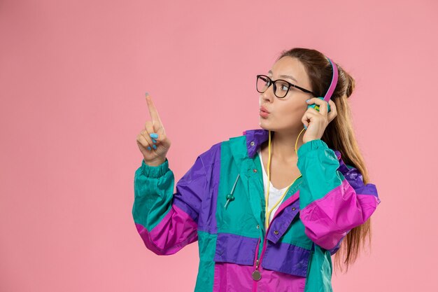 Vista frontal joven mujer atractiva en abrigo de color camiseta blanca escuchando música sobre fondo rosa