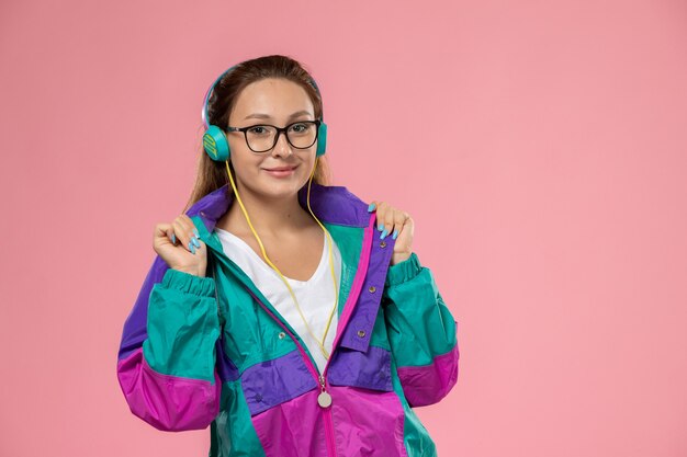 Vista frontal joven mujer atractiva en abrigo de color camiseta blanca escuchando música sobre fondo rosa