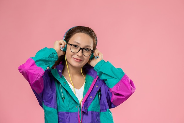 Vista frontal joven mujer atractiva en abrigo de color camiseta blanca escuchando música sobre fondo rosa