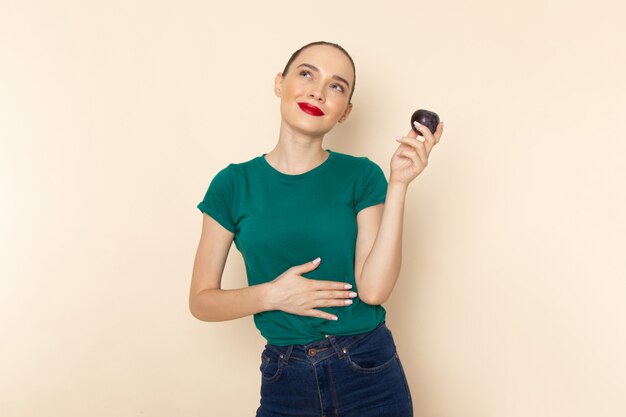 Vista frontal joven mujer atractiva en camisa verde oscuro sosteniendo ciruela y sonriendo en beige