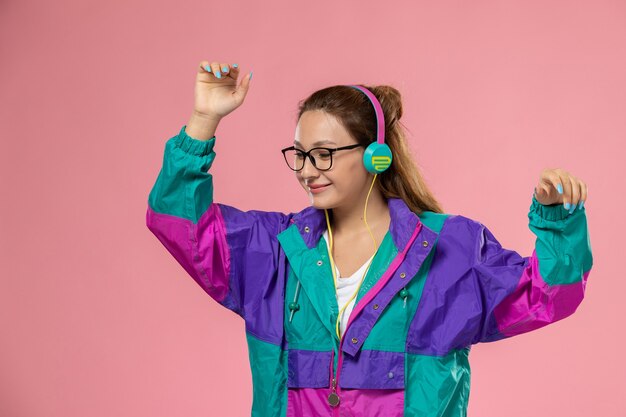 Vista frontal joven mujer atractiva en camiseta blanca abrigo de color bailando escuchando música sobre fondo rosa