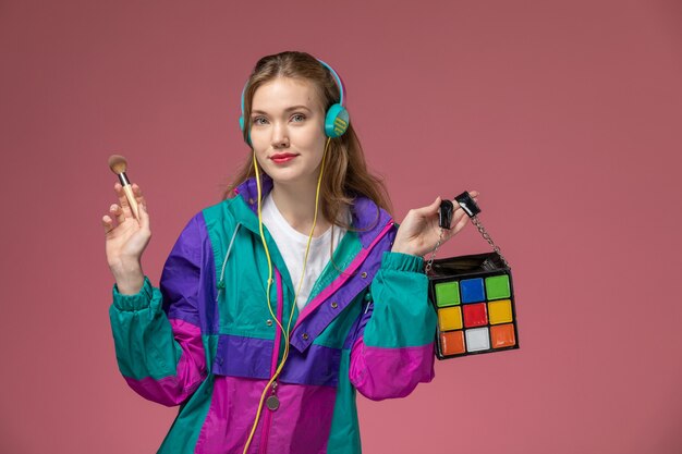 Vista frontal joven mujer atractiva en camiseta blanca abrigo de color escuchando música con auriculares en la pared rosa modelo pose femenina foto en color