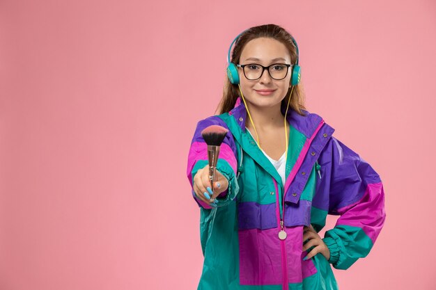 Vista frontal joven mujer atractiva en camiseta blanca abrigo de color escuchando música y haciendo maquillaje en el fondo rosa