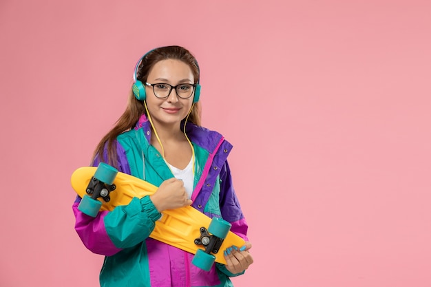 Vista frontal joven mujer atractiva en camiseta blanca abrigo de color escuchando música con una leve sonrisa en el escritorio rosa