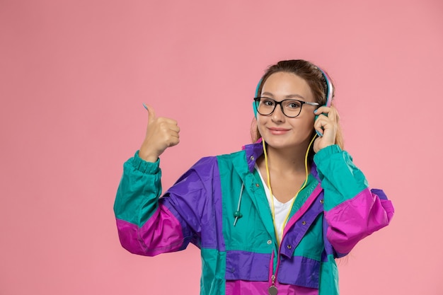 Vista frontal joven mujer atractiva en camiseta blanca y abrigo de color escuchando música y sonriendo sobre el fondo rosa