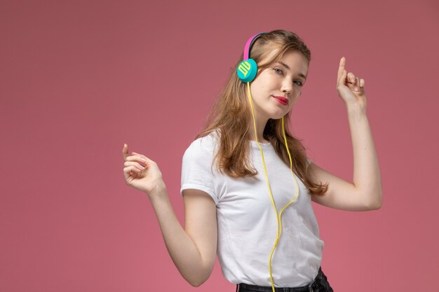 Vista frontal joven mujer atractiva en camiseta blanca bailando y escuchando música en la pared de color rosa oscuro modelo color mujer joven