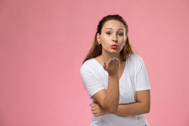 Vista frontal joven mujer atractiva en camiseta blanca enviando besos al aire sobre el fondo rosa