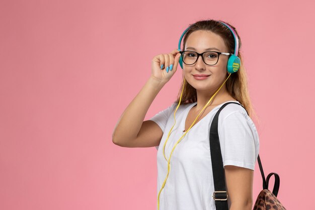 Vista frontal joven mujer atractiva en camiseta blanca escuchando música con auriculares sonriendo en el escritorio rosa