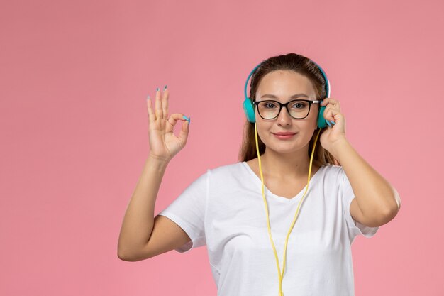 Vista frontal joven mujer atractiva en camiseta blanca escuchando música con auriculares y sonrisa en el fondo rosa