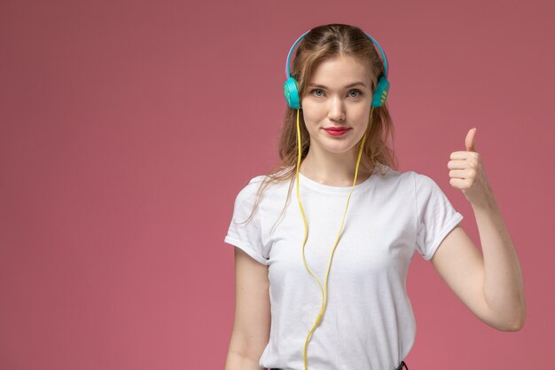 Vista frontal joven mujer atractiva en camiseta blanca escuchando música con una ligera sonrisa en el modelo de escritorio rosa color mujer joven