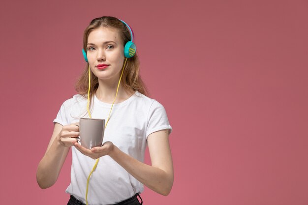 Vista frontal joven mujer atractiva en camiseta blanca escuchando música sosteniendo la taza en la pared rosa modelo color hembra joven