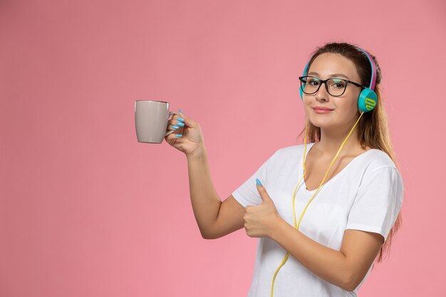 Vista frontal joven mujer atractiva en camiseta blanca escuchando música sosteniendo una taza sonriendo sobre el fondo rosa