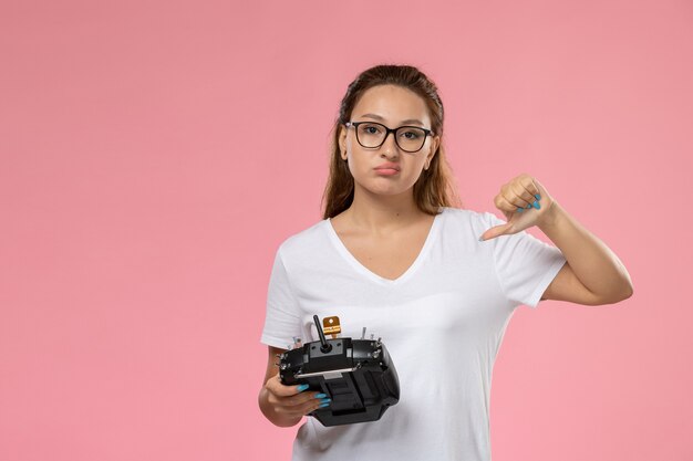 Vista frontal joven mujer atractiva en camiseta blanca con expresión de disgusto sosteniendo el control remoto sobre el fondo rosa