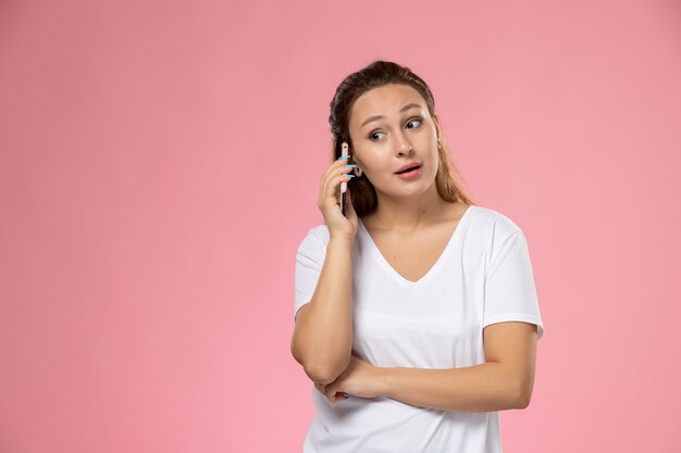 Vista frontal joven mujer atractiva en camiseta blanca hablando por teléfono sobre el fondo rosa