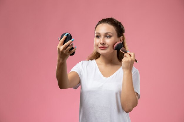 Vista frontal joven mujer atractiva en camiseta blanca haciendo un maquillaje con una leve sonrisa en el fondo rosa