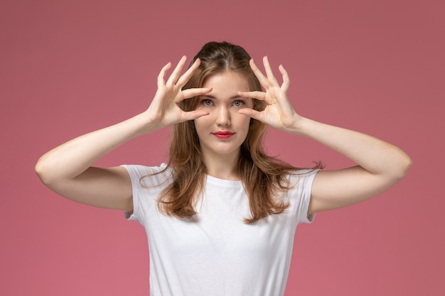 Vista frontal joven mujer atractiva en camiseta blanca mostrando sus ojos posando en la pared rosa modelo pose femenina fotografía en color