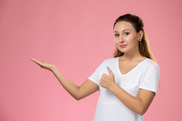 Vista frontal joven mujer atractiva en camiseta blanca de pie y posando sobre fondo rosa