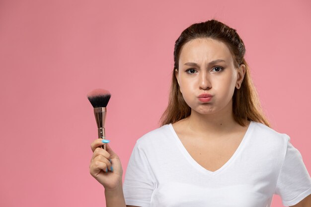 Vista frontal joven mujer atractiva en camiseta blanca con pincel de maquillaje sobre fondo rosa