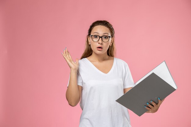 Vista frontal joven mujer atractiva en camiseta blanca posando con archivo gris sobre fondo rosa