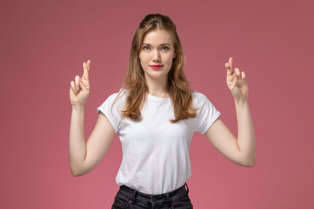 Vista frontal joven mujer atractiva en camiseta blanca posando con los dedos cruzados y sonrisa en la pared rosa modelo pose femenina foto en color