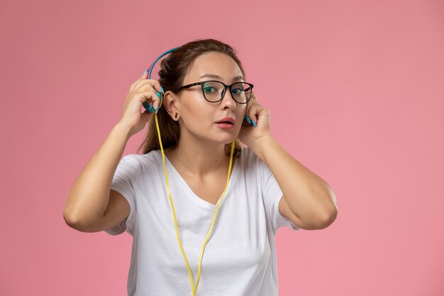 Vista frontal joven mujer atractiva en camiseta blanca posando escuchando música con auriculares en el fondo rosa