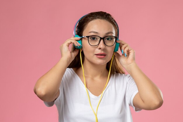 Vista frontal joven mujer atractiva en camiseta blanca posando y escuchando música a través de auriculares en el fondo rosa