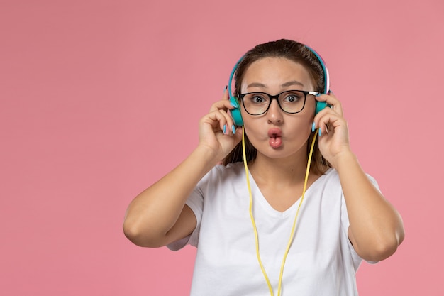 Vista frontal joven mujer atractiva en camiseta blanca posando y escuchando música a través de auriculares en el fondo rosa